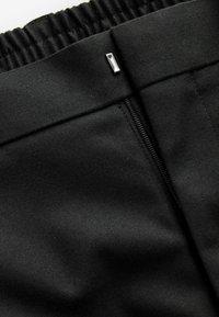 Pantaloni din țesătură neagră, cu un finisaj texturat, talie elastică, închidere cu fermoar și accesorii metalice la talie.