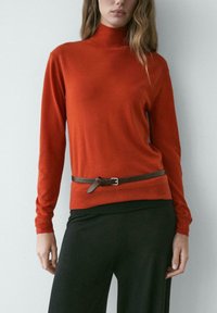 Frau trägt einen enganliegenden Pullover mit Rollkragen in verbranntem Orange, einen schmalen braunen Gürtel und lockere schwarze Hosen, steht vor einer schlichten Wand.