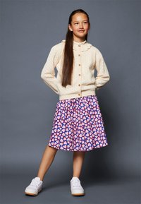 Cardigan in crema a maglia cable con colletto arrotondato, abbinato a una gonna colorata e stampata con cuori rosa su uno sfondo blu. Sneakers bianche.