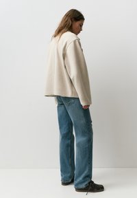 Kobieta o długich włosach ubrana w beżową, oversize'ową kurtkę, niebieskie proste dżinsy i ciemnobrązowe buty, stojąca na tle jasnego, jednolitego tła.