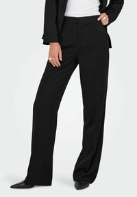 Zwarte, op maat gemaakte wijde pantalon met een gladde afwerking en voorplooien, gecombineerd met puntige zwarte schoenen. Schoon ontwerp, geen zichtbare patronen of accenten.