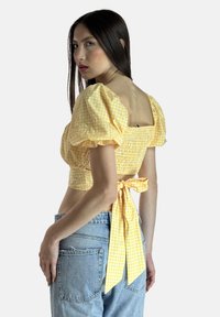 Gul gingham-vest med pufærmer, smockdetaljer foran, åben ryg og bindebånd ved taljen. Bruges med blå denimjeans.