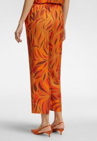 Pantaloni arancioni con un motivo floreale in sfumature di rosso e marrone. Vita elastica e lunghezza croppata. Indossati con tacchi arancioni.