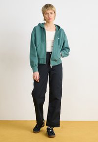 Grønn bomulls hettegenser med glidelås og snorhette, frontlommer, brukt over en hvit tanktop. Kombinert med svarte vide bukser og svarte sneakers.