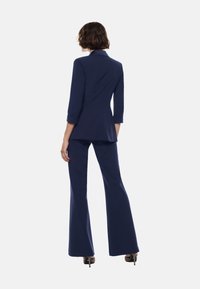 Completo da uomo in blu marina, caratterizzato da un blazer aderente con maniche a tre quarti e pantaloni svasati; tessuto morbido e dettagli di sartoria sottili.