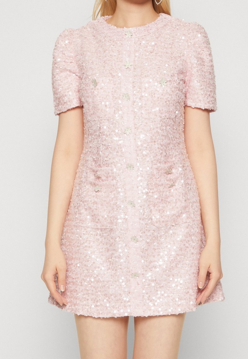 Femme portant une robe texturée rose clair à manches courtes, ornée de sequins, avec des boutons devant et deux poches poitrine ainsi que deux poches sur les hanches.