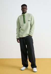 Lys grønn fleecegenser med høy krage, knapper og brystlomme; kombinert med løse sorte jeans og hvite joggesko med grønne detaljer.