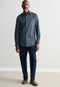 Camisa de vestir de patrón navy con mangas largas, frente abotonado y corte ajustado, combinada con pantalones oscuros y zapatos de ante marrón.
