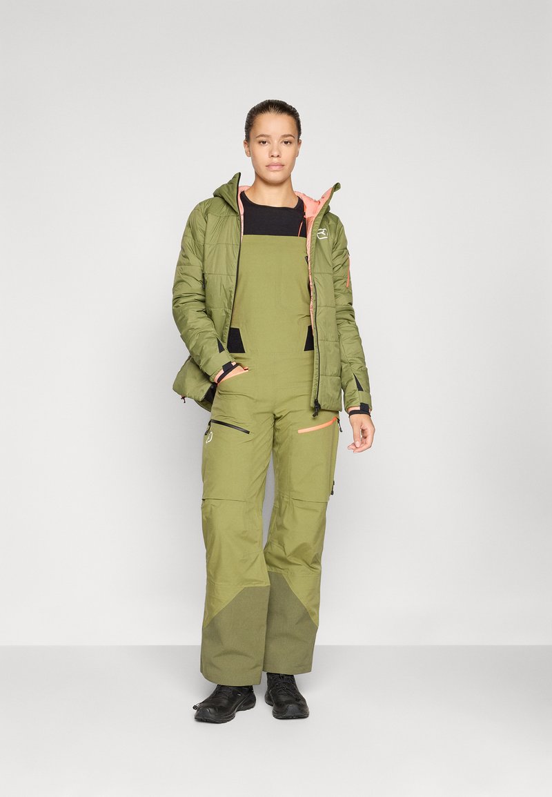 Grüne isolierte Jacke mit Kapuze, kombiniert mit grünen Latzhosen mit schwarzen Akzenten und Reißverschlüssen. Schwarze Schuhe runden das Outfit ab.