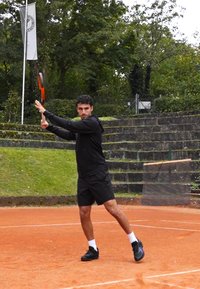 Egy fekete sportöltözetbe öltözött férfi egy narancssárga keretes teniszütőt lenget egy salakos pályán, zöld növényzet és kő lépcsők környezetében.