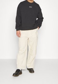 Personne portant un sweat-shirt noir à manches longues avec le texte "WRSTBHVR", un pantalon cargo blanc ample, et des chaussures en cuir noir avec des chaussettes blanches.