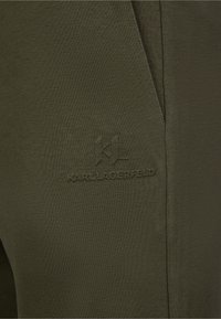 Olivgrön tyg med en slät textur har en präglad "KL"-logotyp. Designen inkluderar en subtil sömdetalj på sidan.