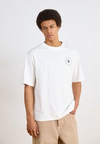 Hvit bomulls t-skjorte med korte ermer, rund hals, og en svart logo trykket på venstre side. Brukt med beige bukser.