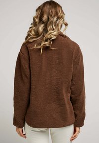 Veste en polaire marron et doux avec des manches longues, une coupe décontractée et un col. Texture douce, sans fermetures visibles et un ourlet arrondi.
