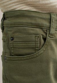 Olijfgroene broek met een voorzak en zwarte knoopdetail, gestikte naden en een gestructureerde stof. De tailleband is zichtbaar aan de bovenkant.