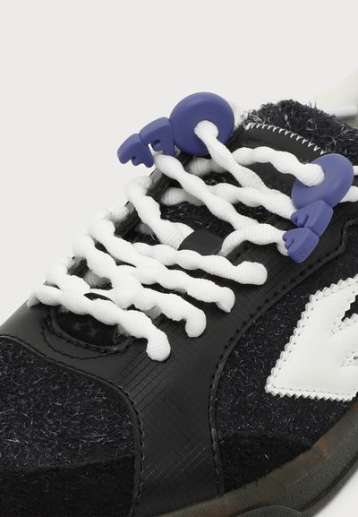 Baskets noires avec des lacets élastiques blancs torsadés, maintenus par des attaches en plastique bleu en forme de disques ronds et de petites mains.