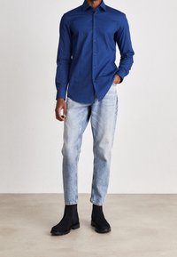 Uomo che indossa una camicia button-up blu aderente, jeans azzurri chiari e stivali neri alla caviglia, in piedi con una mano in tasca.