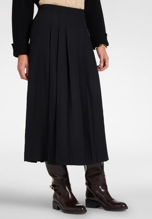 Donna con gonna midi plissettata nera, maglione nero, top beige e stivaletti in pelle marrone scuro con dettaglio fibbia dorata.