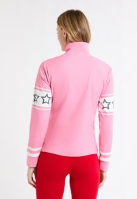 Różowy top z długim rękawem i wysokim kołnierzem, zdobiony białymi paskami i wzorami gwiazdek na rękawach. Gładka, elastyczna tkanina. Czerwone legginsy na dole.