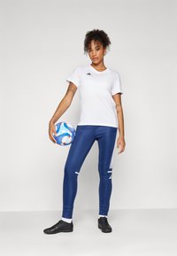 Baltas īsām piedurknēm t-krekls ar melnu logotipu, zili sporta legingi ar baltām svītrām, melnas futbola kurpes, tur rokās zilu un baltu futbola bumbu.
