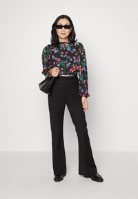 Blusa preta de mangas longas com padrões multicoloridos, combinada com calças flare pretas de cintura alta e sapatos de couro pretos. Segura uma bolsa preta.