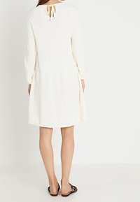 Langärmliges weißes Kleid mit einem gerippten Muster, rundem Ausschnitt und einer Bindeschließe im Rücken. Leichter Stoff und eine ausgestellt geschnittene Silhouette.