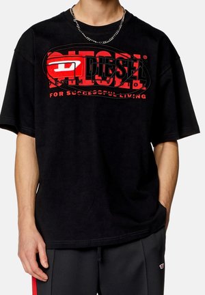 Personne portant un t-shirt Diesel noir avec un logo rouge et blanc et un texte, associé à un pantalon foncé et un collier en chaîne argenté.
