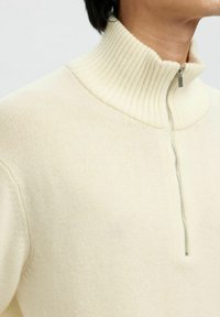 Cremefarbener Pullover mit hohem geripptem Kragen und Viertelreißverschluss. Weiche Stricktextur mit minimalen Nähdetails, die ein sauberes Design bieten.