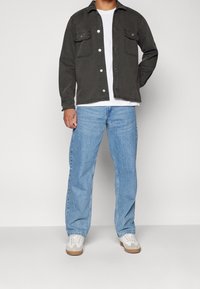 Man som bär en mörk knapp-up jacka över en vit t-shirt, ljusblå jeans och vita sneakers, stående mot en enfärgad bakgrund.