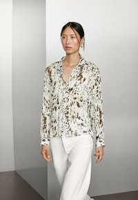 Blouse à manches longues en tissu léger, ornée d'un imprimé abstrait beige et marron, assortie à un pantalon blanc pour un look harmonieux.