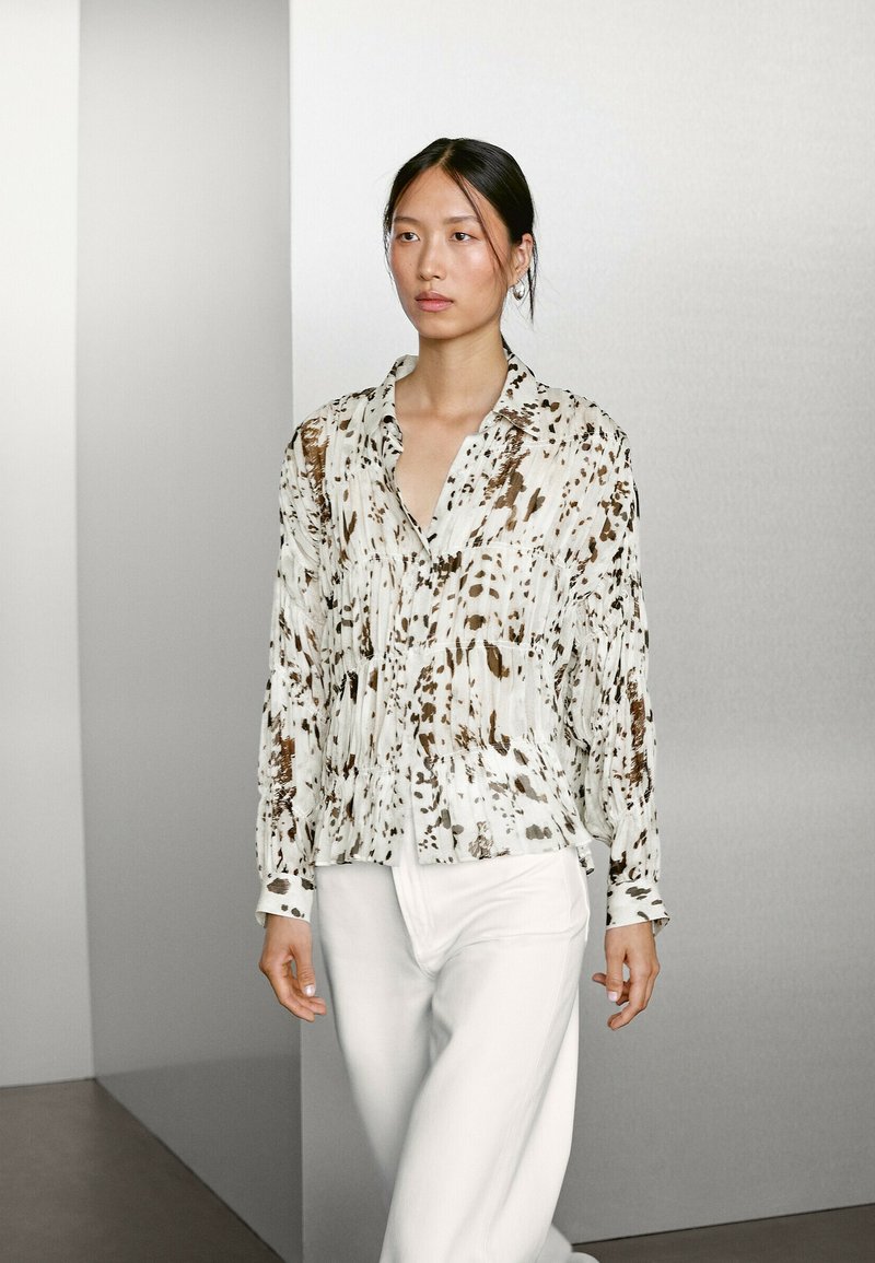 Blouse à manches longues en tissu léger, ornée d'un imprimé abstrait beige et marron, assortie à un pantalon blanc pour un look harmonieux.