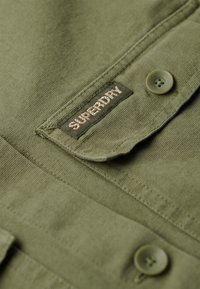 Camisa verde oliva con una tela texturada, que presenta una etiqueta de Superdry y un cierre de botones. Los detalles notables incluyen un botón redondeado.