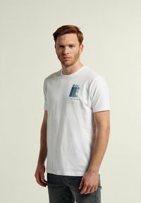 Camiseta blanca de algodón, con un estampado gráfico en turquoise y azul en la zona izquierda del pecho. Manga corta y cuello redondo.
