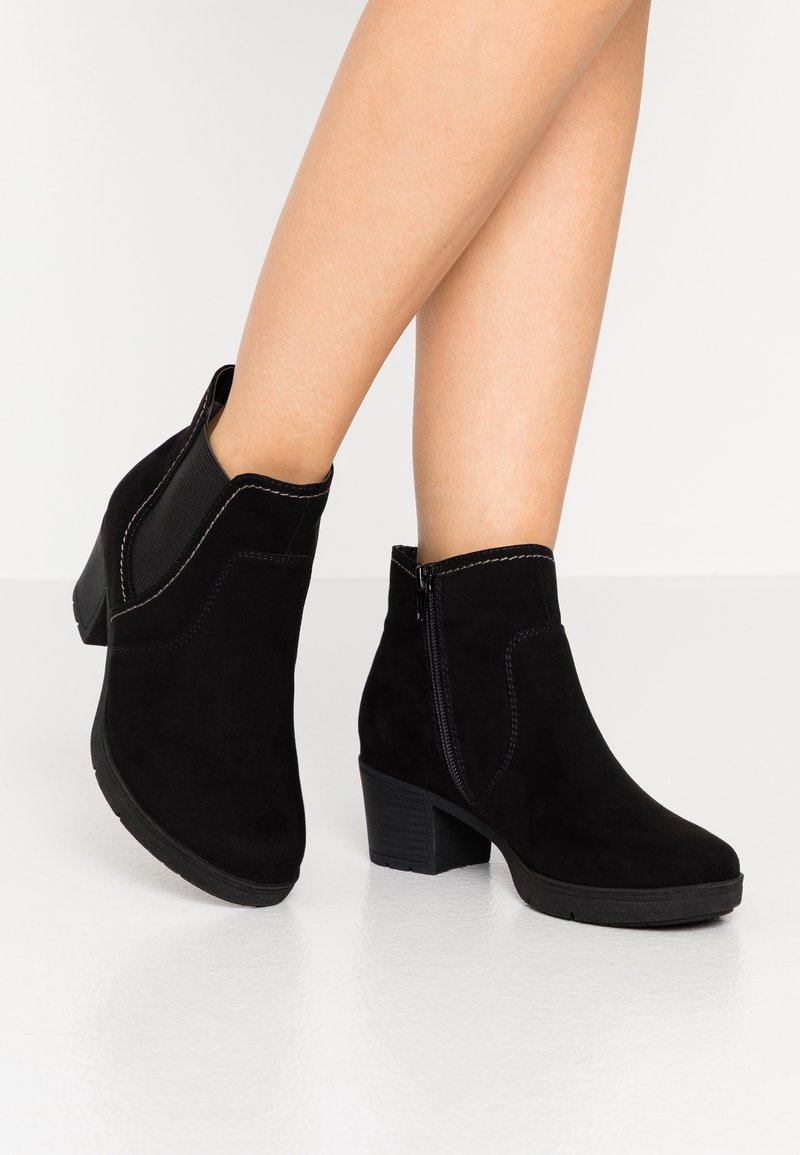 Bottines noires en suède à talons blocs portées sur jambes nues sur fond blanc, montrant une fermeture éclair latérale et un panneau élastique.