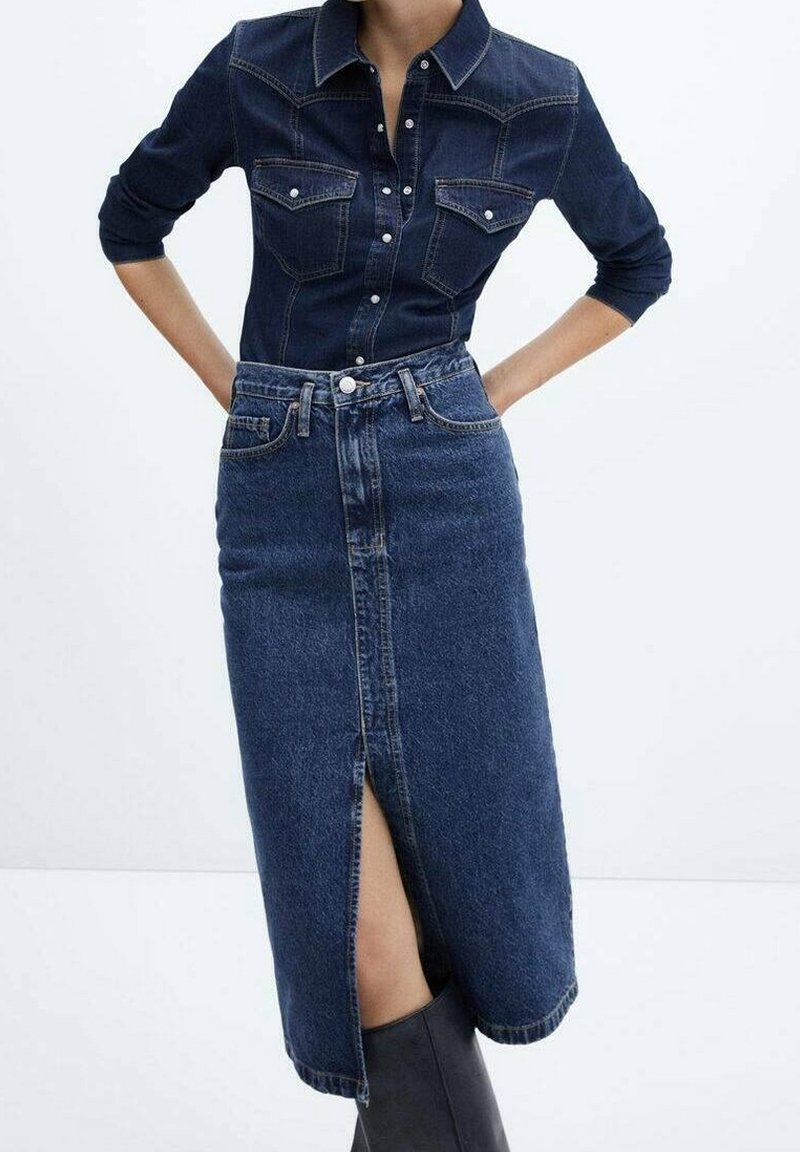 Femme portant une chemise en jean foncé boutonnée et une jupe midi en jean taille haute assortie avec une fente à l'avant, associées à des bottes noires montantes jusqu'aux genoux.