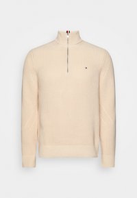 Pull en maille beige avec col zippé à mi-hauteur, poignets et ourlet côtelés, arborant un petit logo de drapeau sur la poitrine gauche.