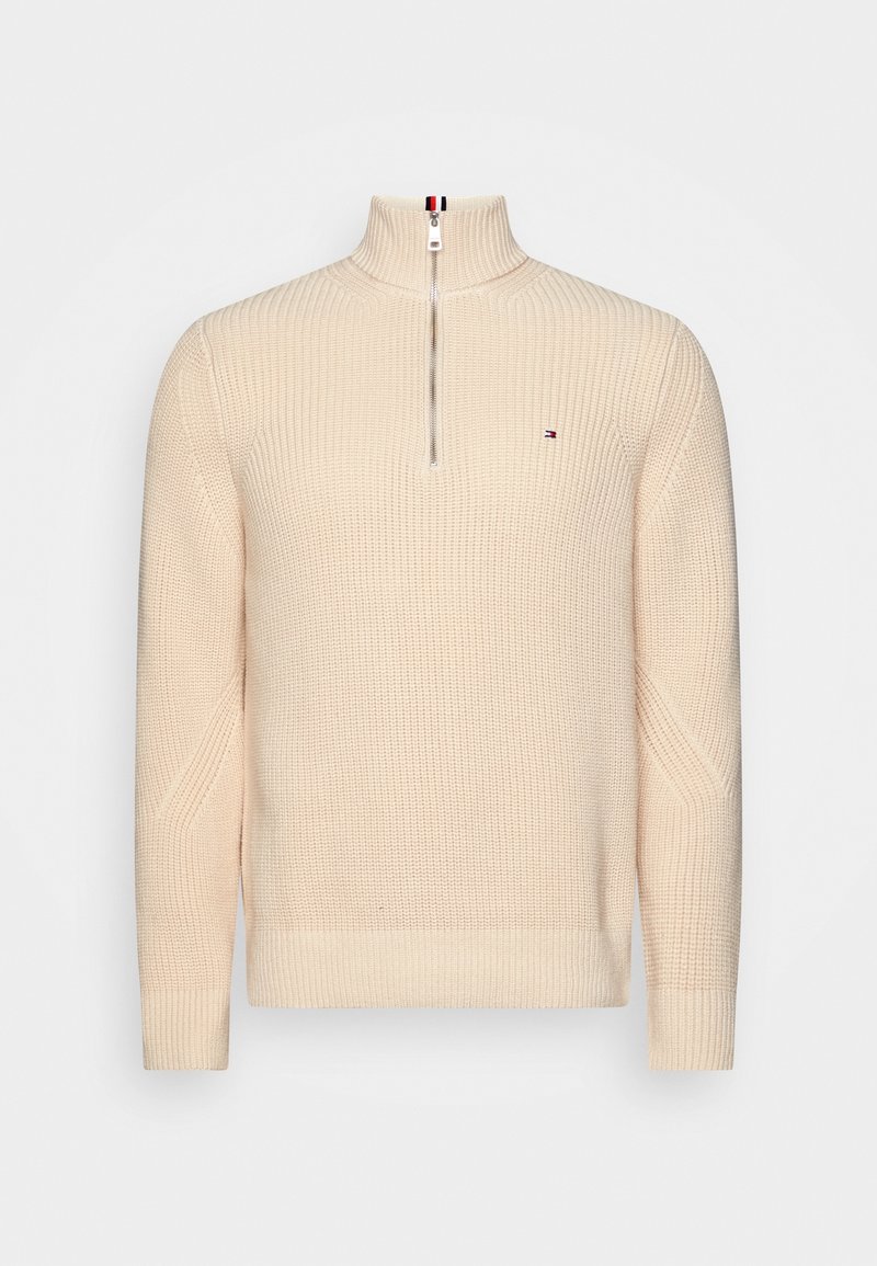 Pull en maille beige avec col zippé à mi-hauteur, poignets et ourlet côtelés, arborant un petit logo de drapeau sur la poitrine gauche.
