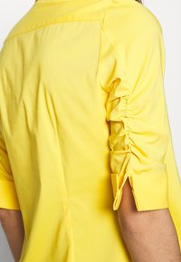 Gul bomullskjorta med rynkade ärmar, knäppta manschetter och en strukturerad design som visar en jämn textur och en livlig färg.