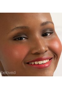 Gros plan d'une personne souriante avec une peau lisse et foncée, portant un maquillage naturel, avec un fard à paupières subtil et des lèvres rouges vives.