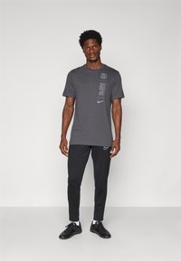 T-shirt en coton gris à manches courtes avec un motif graphique. Associée à un pantalon noir ajusté arborant un petit logo Nike sur la cuisse.