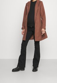 Abrigo marrón de longitud media con capucha, hecho de tela texturizada; combinado con pantalones de campana negros y botas negras de tobillo con suela gruesa.