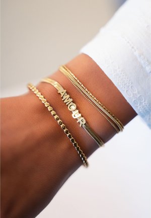 Pulseras de oro apiladas en una muñeca: una con la inscripción "AMOUR", una cadena texturizada, una bola redonda lisa y un diseño de malla.