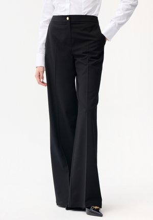 Kvinde iført sorte, vidde bukser med højt talje og plisseringer, en hvid skjorte med knapper og sorte spidse hæle med gulddetaljer.