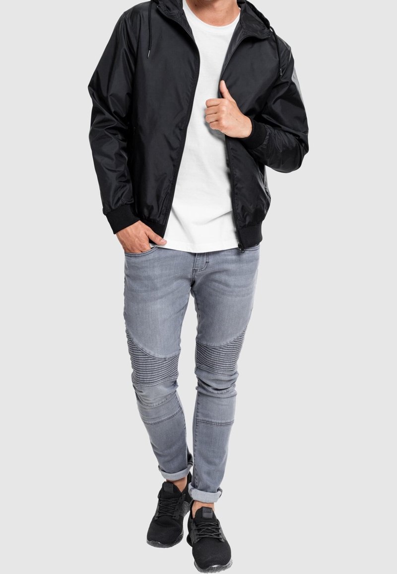 Black zippered jacket with hood, white t-shirt underneath, gray jeans with ribbed accents, and black sneakers. Modern casual outfit.