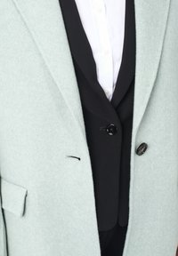 Blazer in verde menta chiaro a spina di pesce con un gilet nero sottostante, caratterizzato da un bottone nero e un piccolo dettaglio di cucitura nera. Camicia bianca visibile.