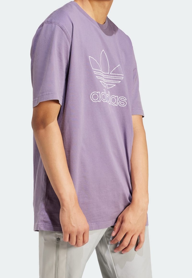 En person som bär en lös lavendelfärgad Adidas t-shirt med en vit logga, kombinerad med ljusgrå byxor med sidofickor med dragkedja.