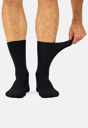 Person trägt schwarze Socken, rechte Hand zieht den oberen Rand der rechten Socke vom Bein weg.