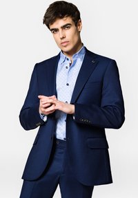 Granatowa marynarka o teksturowanym wykończeniu, połączona z jasnoniebieską koszulą w mały wzór kropek. Model stoi z dłońmi złożonymi.