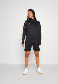 Zwarte sweatshirt met capuchon en ritszakken, met een gouden logo en accenten. Gekoog met zwarte shorts en witte sneakers.