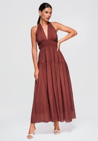 Femme aux cheveux foncés en queue-de-cheval basse portant une robe maxi halter marron rouille sans manches et des talons blancs à bouts ouverts, posant devant un fond uni.